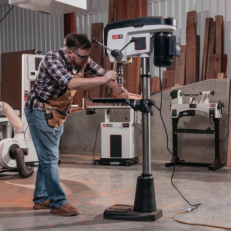 A man wearing safety glasses, a plaid shirt, and an apron is using a pedestal drill in a workshop. The workspace features other woodworking equipment, and he is carefully drilling into a piece of wood.