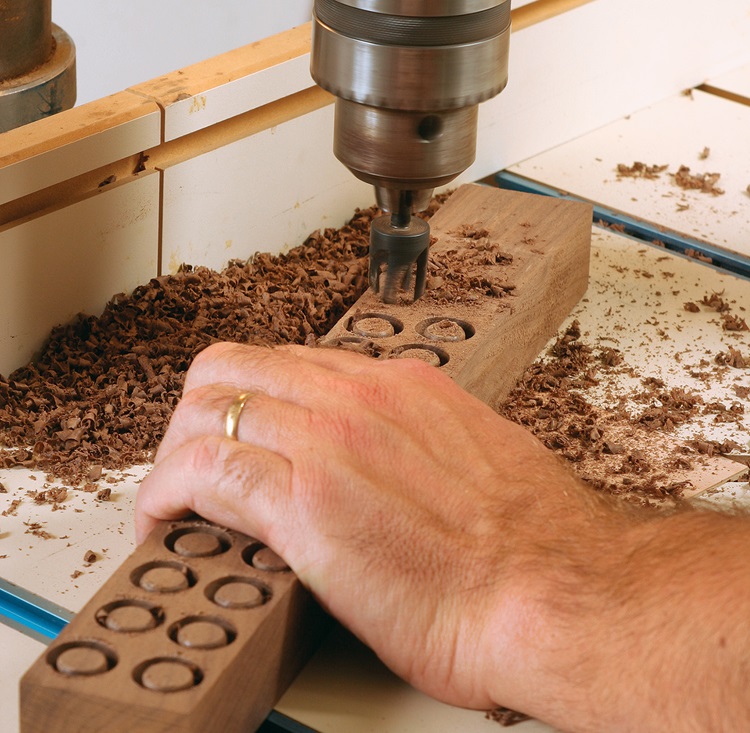 Close-up of a pedestal drill in action, creating evenly spaced circular holes in a wooden block. Shavings from the drilled wood are scattered around, while a hand steadies the block.