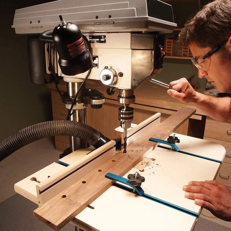 A focused woodworker adjusts a piece of wood clamped on the table of a pedestal drill, ready to make precise vertical holes. The machine is equipped with guides and clamps to ensure accuracy.